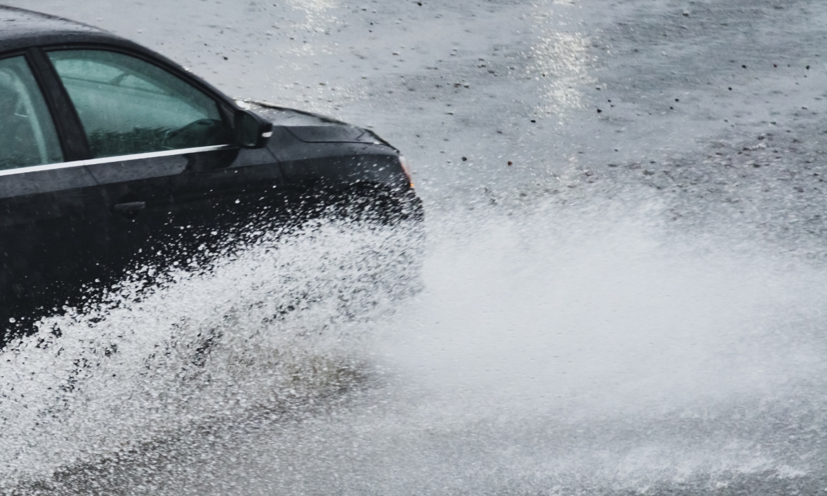 a car driving through a large puddle in the rain, the puddle splashing up very high and making a wave of sorts